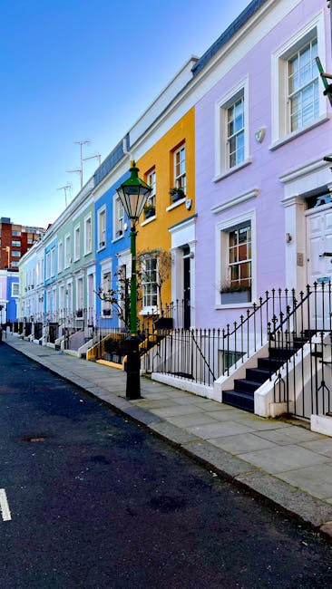 A row of colorful terraced houses painted in pastel shades of blue, yellow, and purple, situated along a city street with a black iron fence and steps leading up to each property's front door. The pav